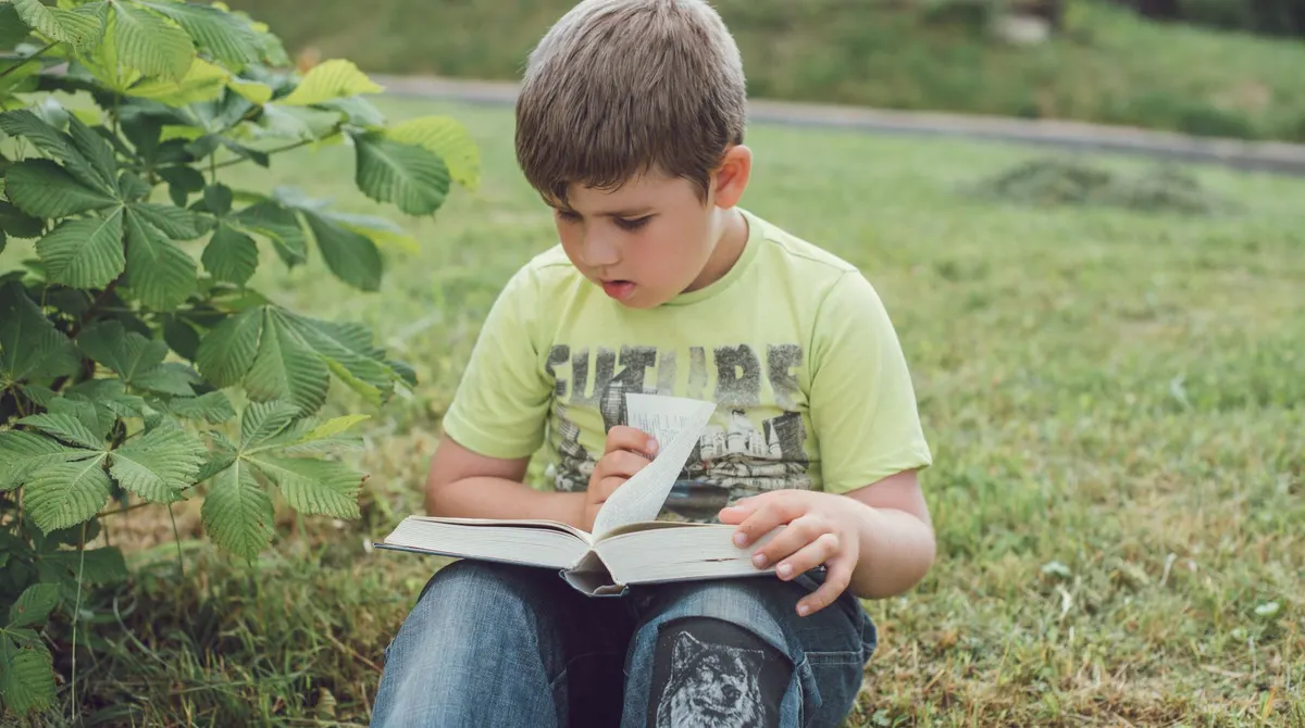 A boy sitting on the grass, turning the page of a thick book resting on his knees